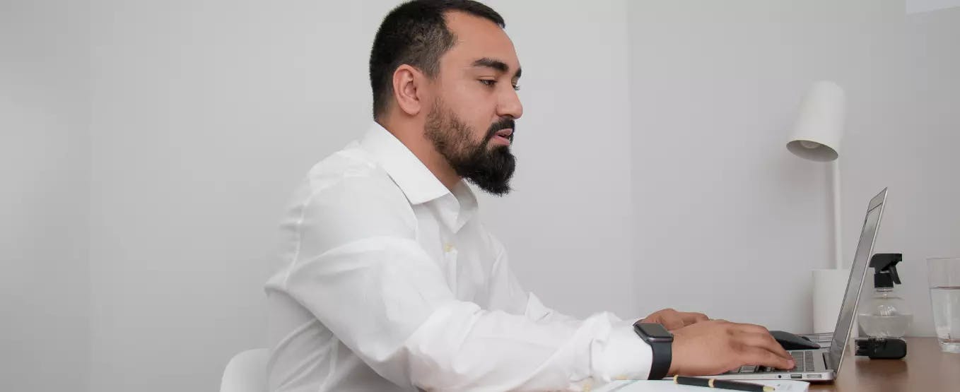 Homem sentado em frente a uma mesa mexendo em um notebook. Ele usa uma camisa social e um relógio em seu pulso.